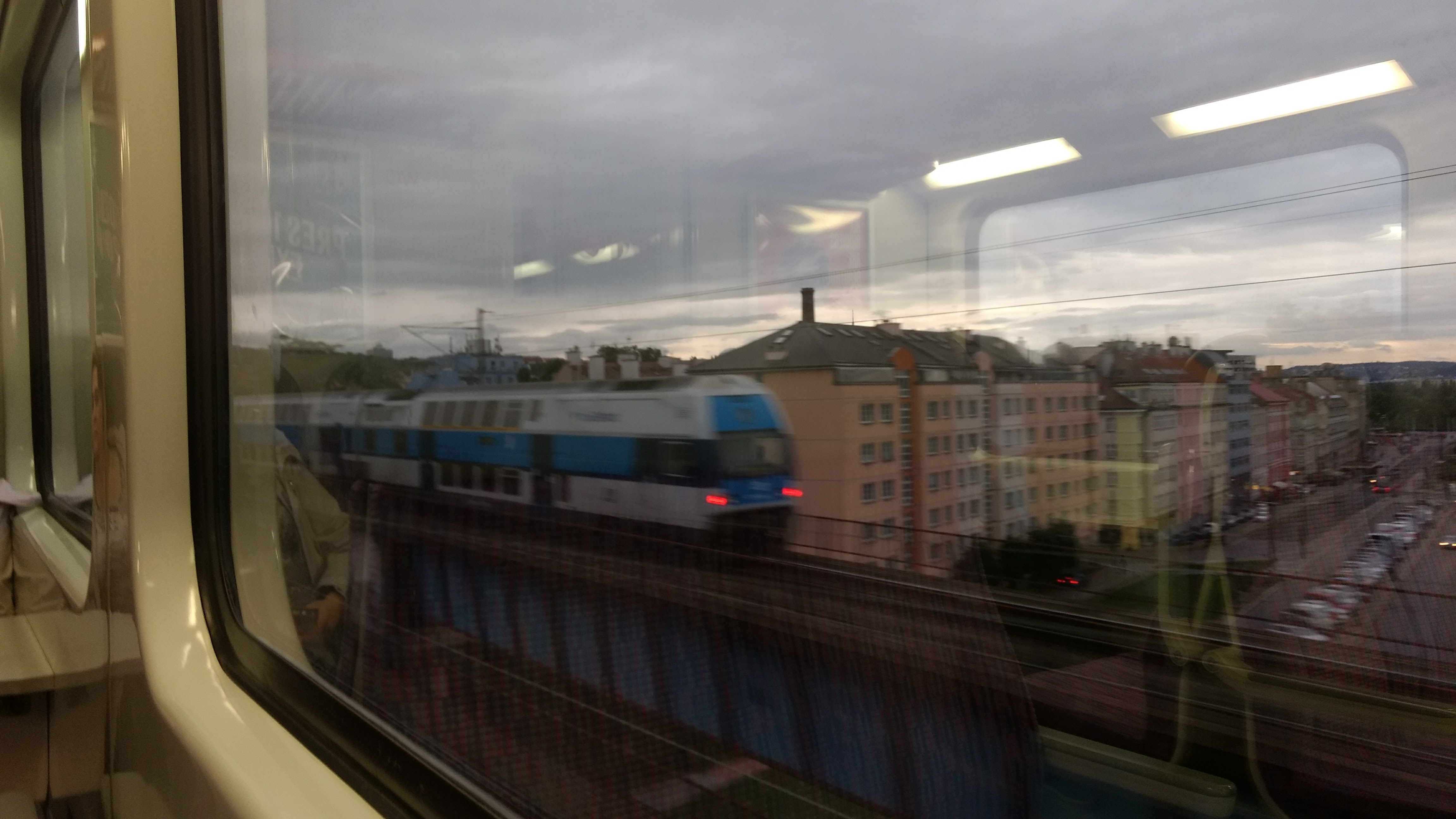 471 class EMU on a bridge over Na Žertvách street, pictured through a window of another train passing on the other track.
